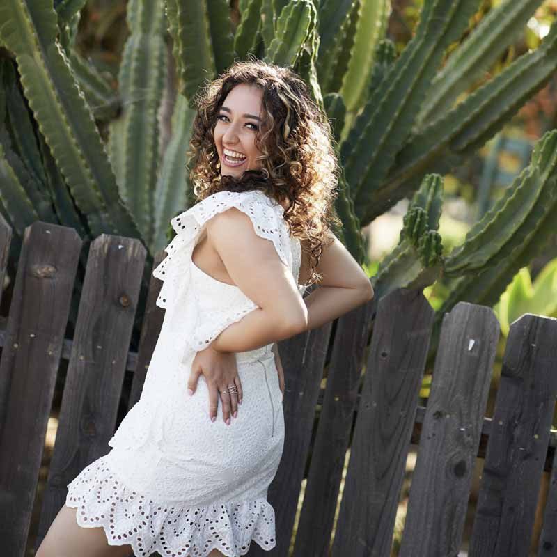 A woman with curly hair wearing a white dress stands smiling by a wooden fence, with large green cacti in the background, creating a joyful, sunny scene.