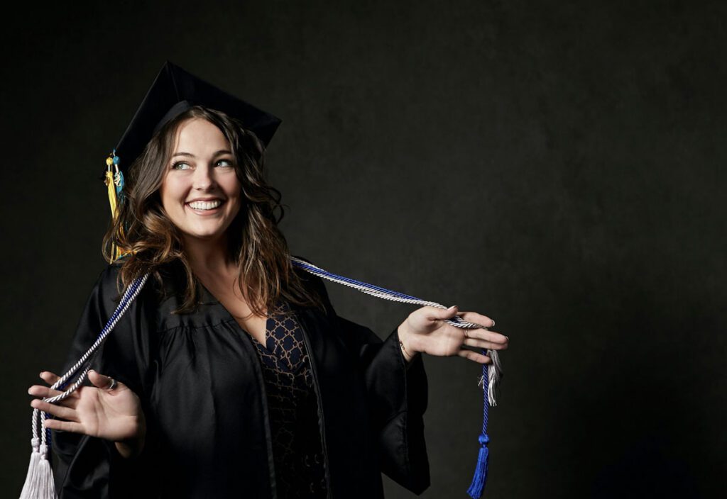 Smiling graduate in a cap and gown, holding blue and white cords against a dark background, conveying joy and achievement.