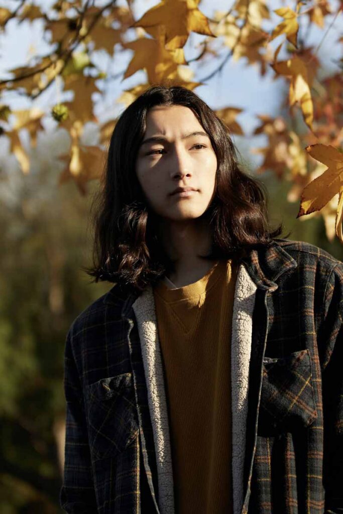 A person with long hair gazes thoughtfully under an autumn tree with golden leaves. They're wearing a plaid jacket, conveying a serene, contemplative mood.