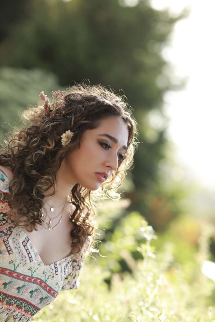 A woman with curly hair adorned with flowers gazes thoughtfully downward in a sunlit garden, wearing a patterned dress. The mood is serene and contemplative.