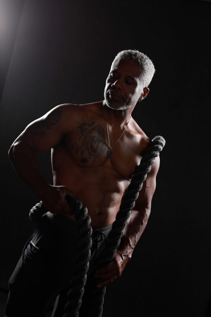 A muscular man exercises with heavy ropes in a dimly lit gym. He is shirtless, revealing tattoos, and expresses focus and determination.