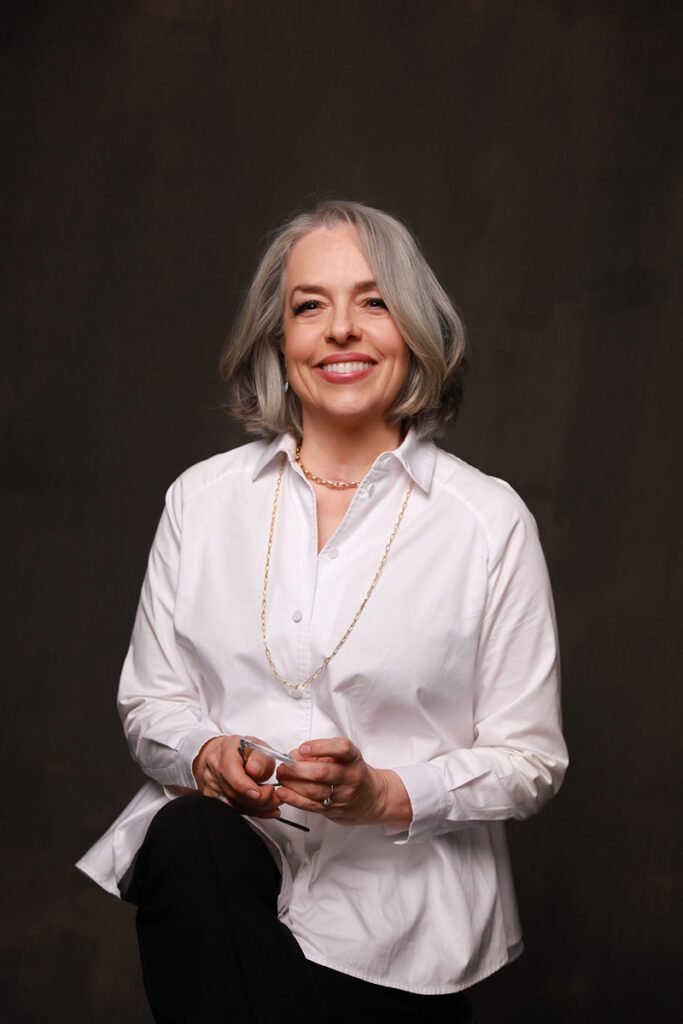 A woman with gray hair smiles warmly, wearing a white blouse and pearl necklace. She holds glasses, seated against a dark background, exuding confidence and elegance.